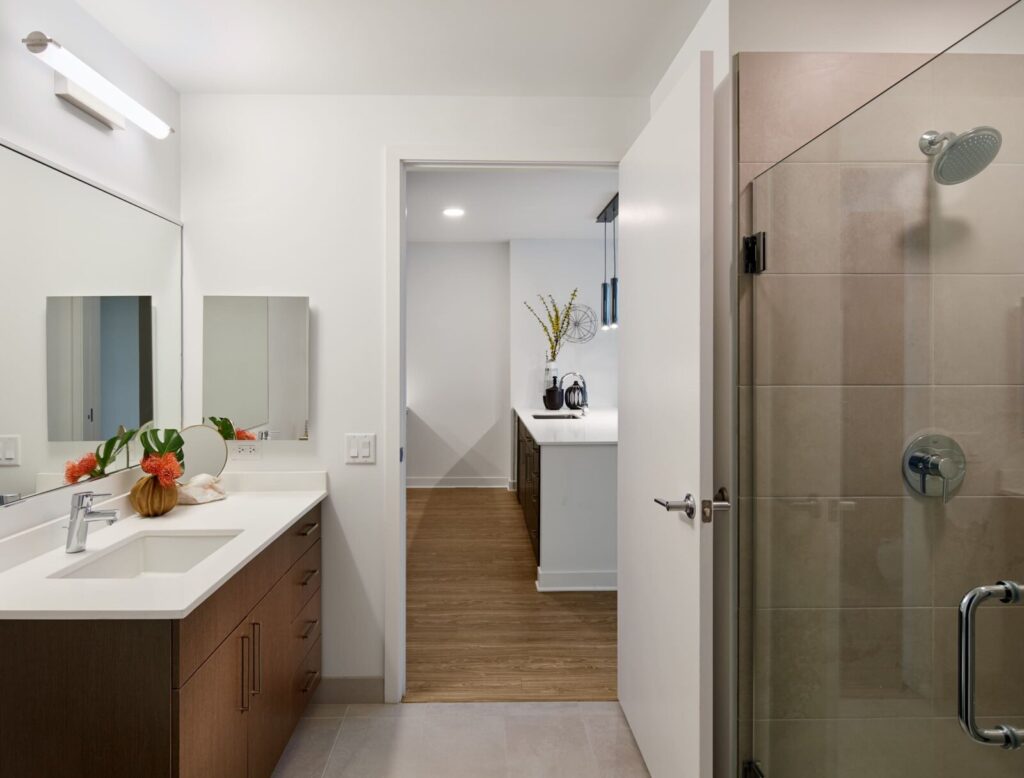 Modern bathroom with a glass shower, wood vanity, and a large mirror, leading into a hallway with light wood flooring and a view of a kitchen counter with a vase of flowers.
