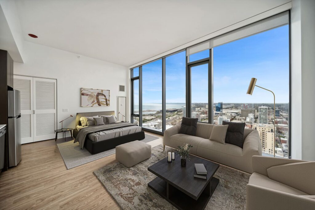 Modern studio apartment with floor-to-ceiling windows, a bed, sofa, coffee table, and cityscape view. Light wood floors and neutral-toned decor create a bright, open space.