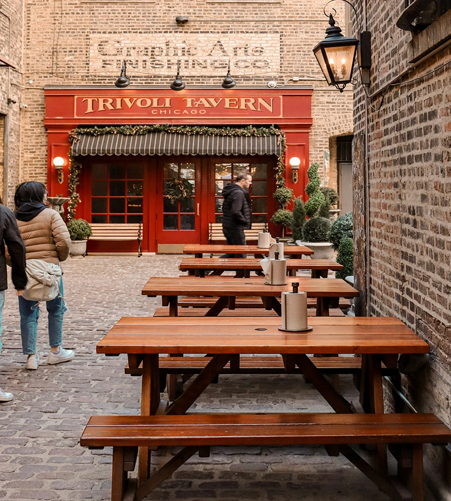 Outdoor seating area with wooden picnic tables leading to a red-bricked facade of Trivoli Tavern in an alley. People are walking towards the tavern entrance.