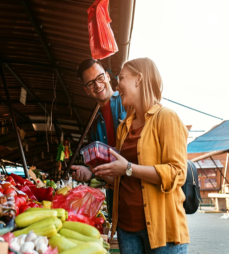 A man and woman smile as they browse produce at an outdoor market stall.