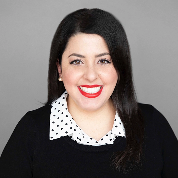 A woman with long dark hair smiles at the camera, wearing a black top and a white collared shirt with black polka dots, against a plain gray background.