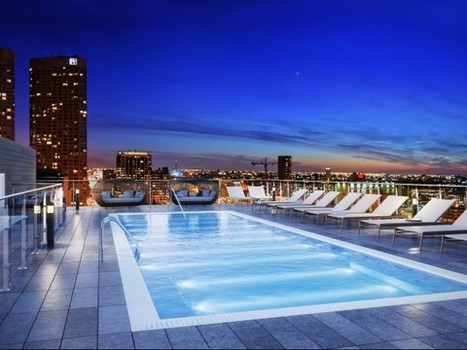 Rooftop swimming pool with lounge chairs, glass railing, and city skyline in the background at dusk.