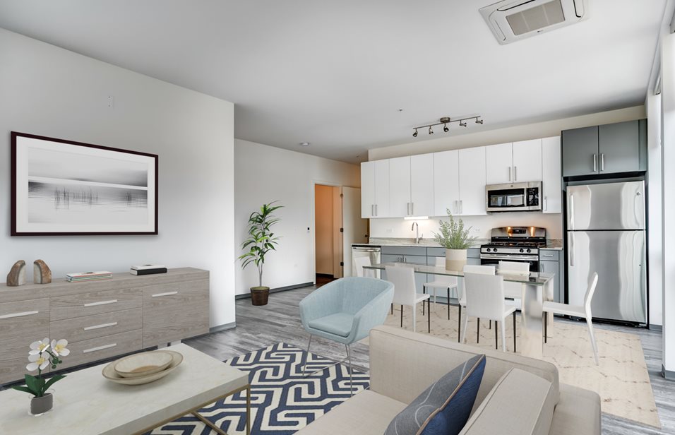 Modern open-plan living area with a kitchen, dining table, and seating space. Features light decor, wood flooring, and a framed picture on the wall. Air conditioner visible on the ceiling.