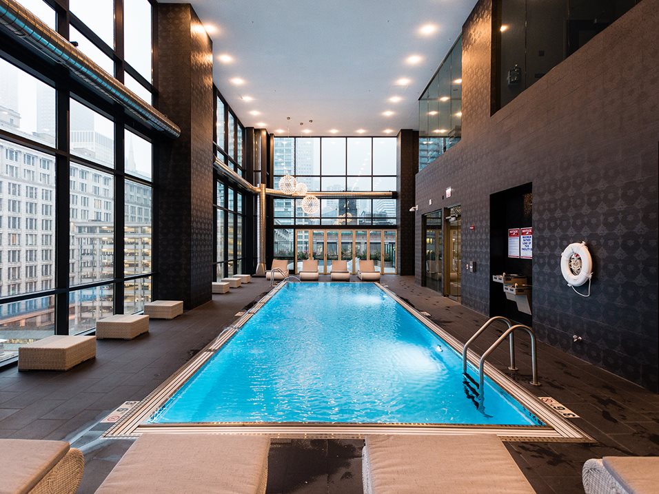 Indoor swimming pool with lounge chairs and large windows offering a city view. The ceiling has recessed lighting, and the walls are dark-toned. Lifebuoys are mounted on the walls.