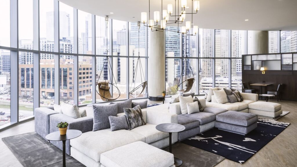 Spacious modern living room with large windows, sectional sofas, hanging chairs, and a cityscape view. A small potted plant and dark tables complement the light decor and open floor plan.