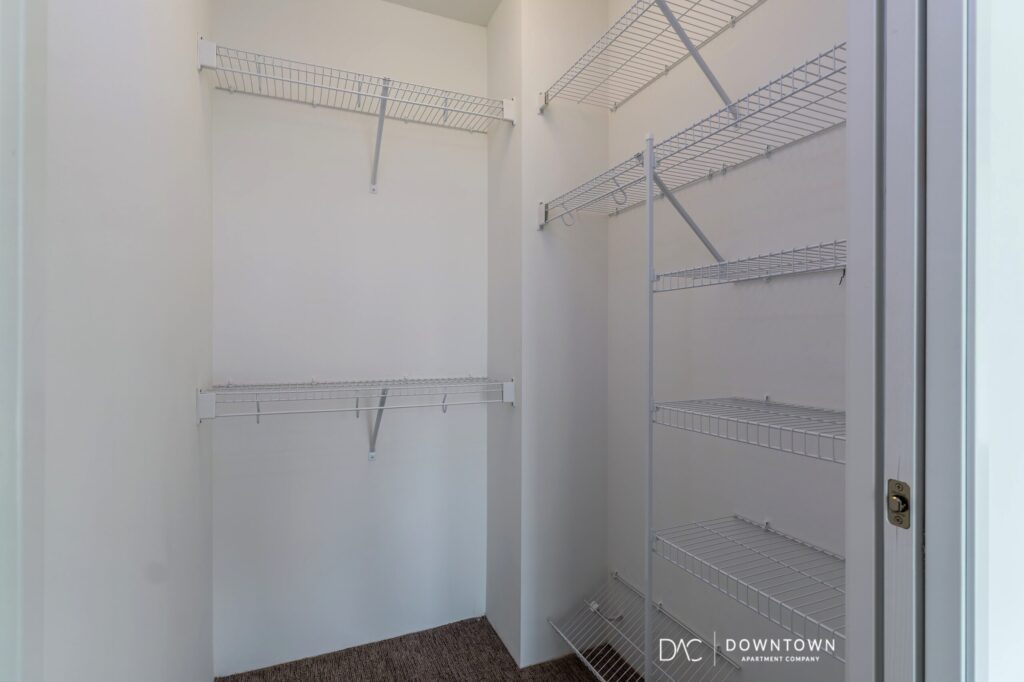 Empty walk-in closet with white wire shelves on two walls and beige carpeted floor.