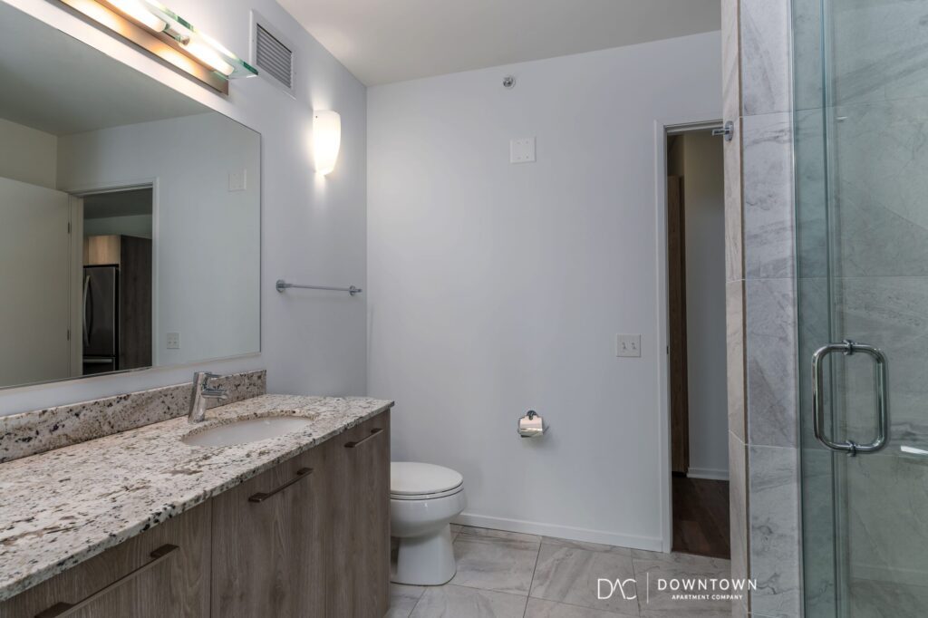 Modern bathroom with a granite countertop, built-in sink, wooden cabinets, toilet, wall-mounted light fixtures, and a glass shower enclosure.