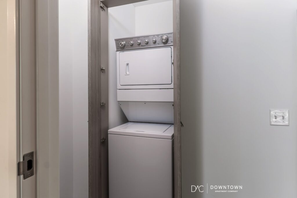 A stacked washer and dryer unit is installed in a small laundry closet with white walls and an open door.