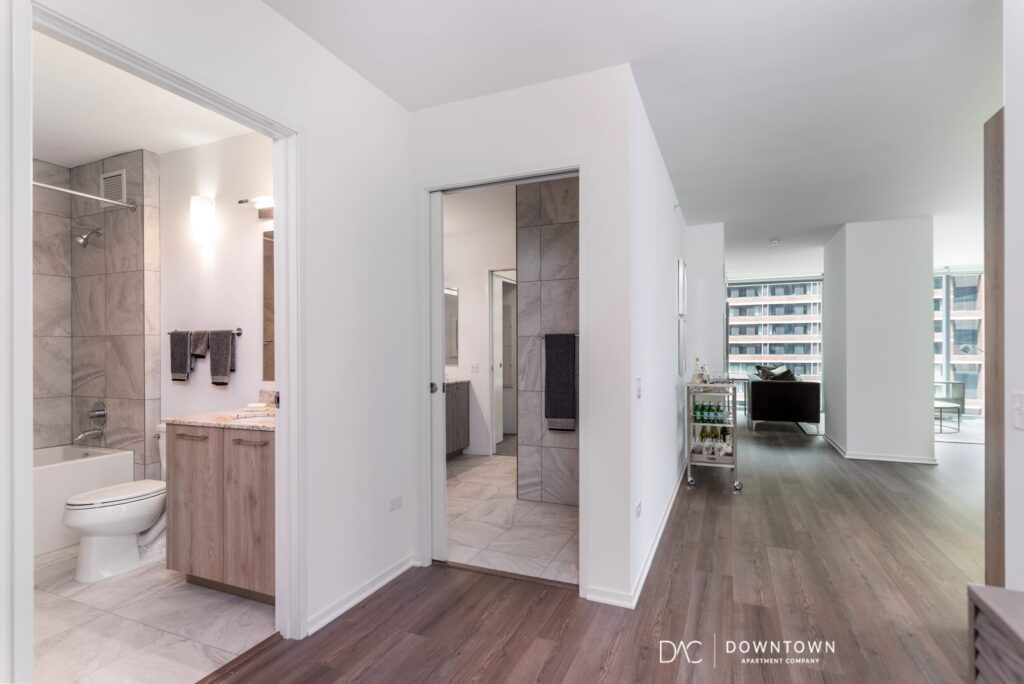 Modern apartment interior with an open hallway view, showing a bathroom, bedroom, and living area with large windows and wood flooring.