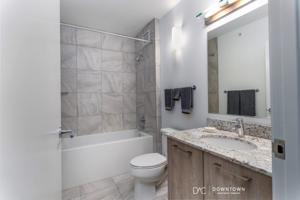 Modern bathroom with a bathtub-shower combo, toilet, granite countertop sink, large mirror, and neutral-colored tiles and towels.
