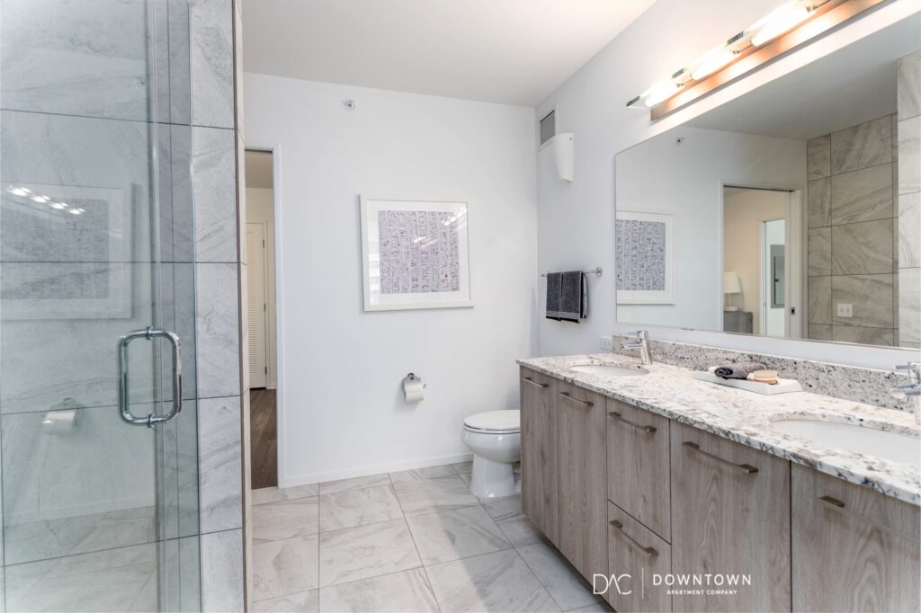 Modern bathroom with glass shower, toilet, double-sink vanity with granite countertop, wall mirror, and tile flooring; neutral color scheme.