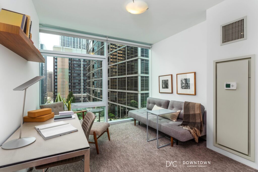 Modern office space with large windows, desk, shelves, sofa, and wall art, overlooking tall city buildings.