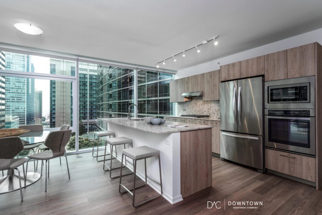 Modern kitchen with light wood cabinets, stainless steel appliances, and a breakfast bar; large windows reveal a cityscape with tall buildings.
