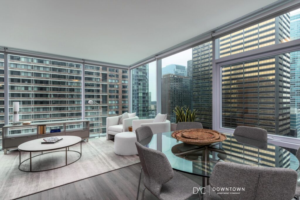 Modern high-rise apartment living room with large windows, gray and white furniture, a glass dining table, and views of surrounding city skyscrapers.