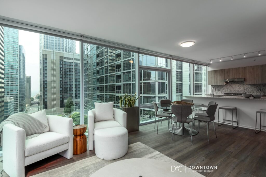 Modern apartment interior with floor-to-ceiling windows, cityscape views, white armchairs, round table with chairs, kitchen area, and contemporary decor.