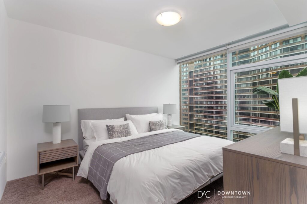Modern bedroom with a neatly made bed, two bedside tables with lamps, a dresser, and large windows with city views.