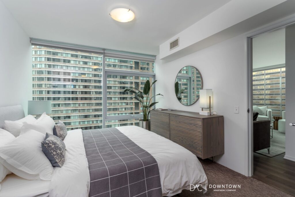 Modern bedroom with a large window, bed with white linens, gray blanket, dresser with round mirror, and a potted plant; city buildings visible outside.