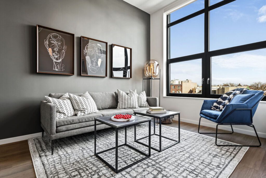 Modern living room with a gray sofa, patterned pillows, blue armchair, two nesting coffee tables, abstract wall art, large window, and a neutral rug on wood flooring.