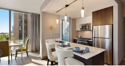 Modern kitchen with an island, bar stools, stainless steel appliances, and a sunlit dining area with city views.