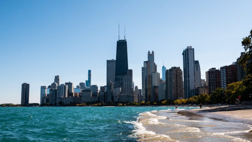 Chicago skyline view from Lake Michigan with high-rise buildings in the background and gentle waves along the shoreline under a clear blue sky.