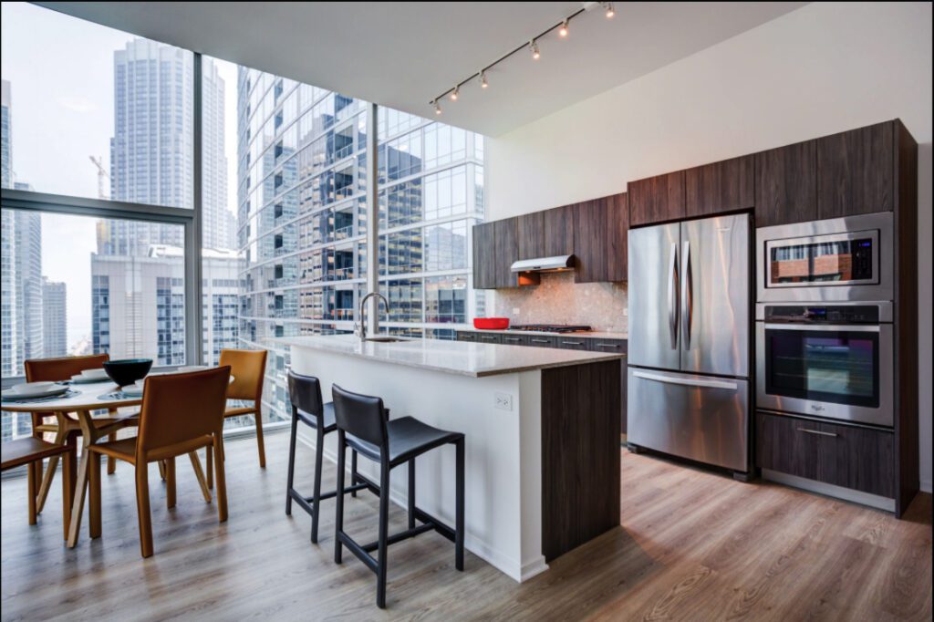 Modern kitchen with stainless steel appliances, wood cabinets, and an island with seating, adjacent to a dining area, set in a high-rise apartment with large windows and city views.