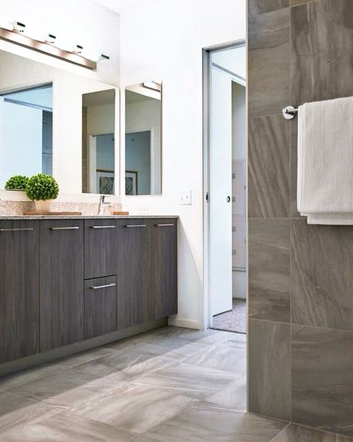 Modern bathroom with gray cabinetry, large mirrors, a plant on the counter, and gray tiled floors and walls; a white towel hangs on the wall near a partially open door.