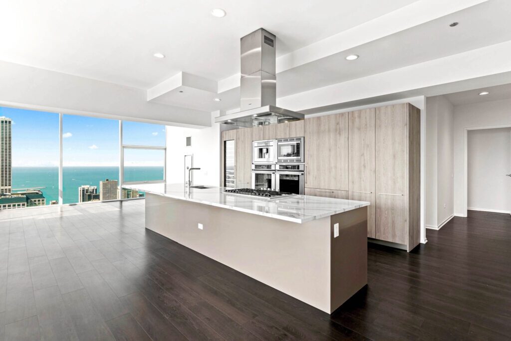 Modern kitchen with a large island, stainless steel appliances, light wood cabinets, dark wood flooring, and floor-to-ceiling windows overlooking a cityscape and water.