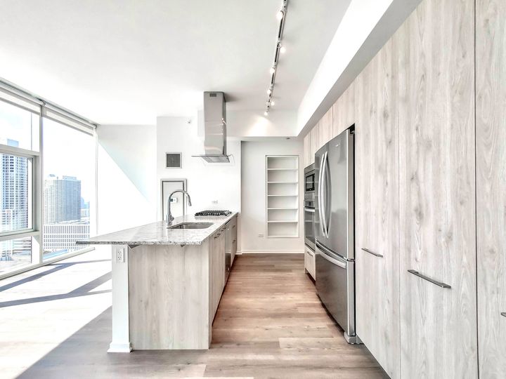 Modern kitchen with light wood cabinets, stainless steel appliances, marble island with sink, track lighting, and large windows offering a city view.