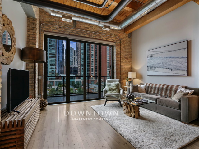 Modern loft apartment with exposed brick walls, wood beams, large windows, and city view. Furnished with a sofa, chair, TV, art, and a round coffee table.