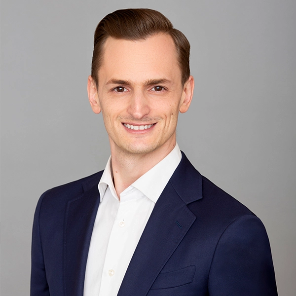 A man in a navy suit jacket and white dress shirt smiles at the camera against a plain gray background.