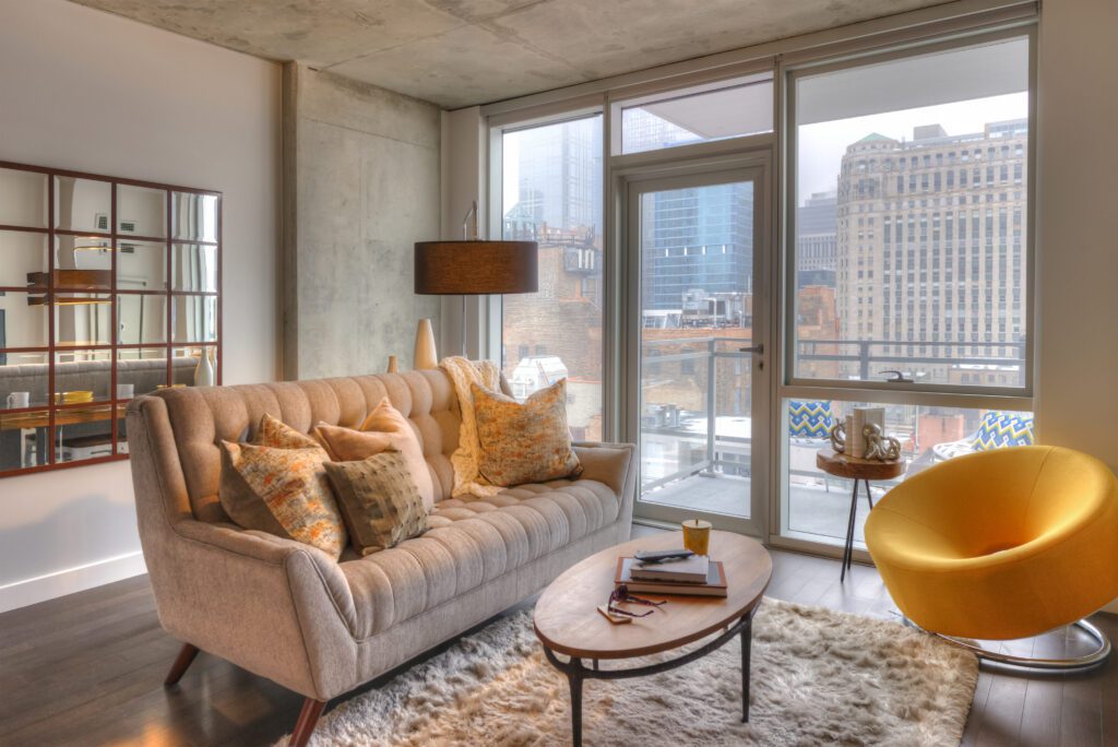 Modern living room with a beige sofa, patterned pillows, a round yellow chair, and a wooden coffee table on a fluffy rug. Large windows reveal a cityscape view.