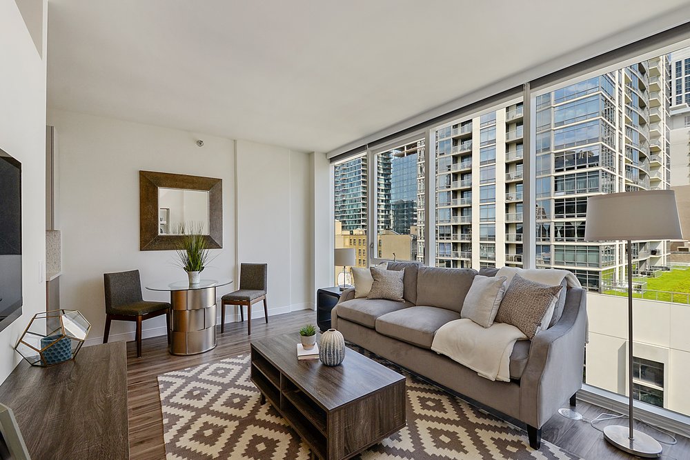 Modern living room with a gray sofa, patterned rug, and a small dining setup with two chairs. Large windows reveal a cityscape with tall buildings and a glimpse of greenery outside.