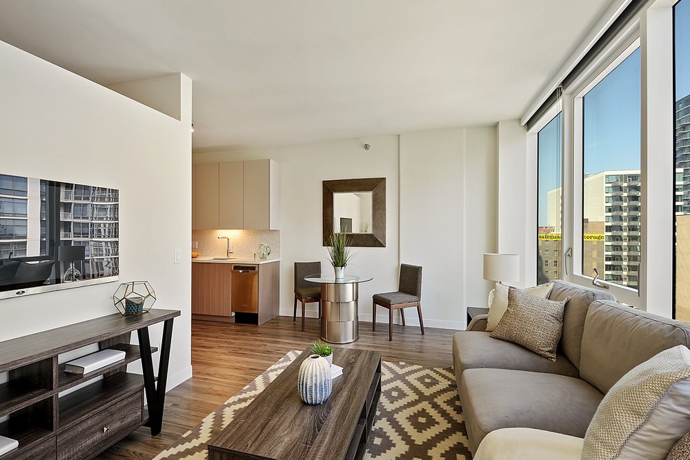 Modern living room with a gray sofa, wooden coffee table, and a geometric rug. Large windows offer a city view. A TV is mounted on a cabinet, and a small kitchen area is visible in the background.