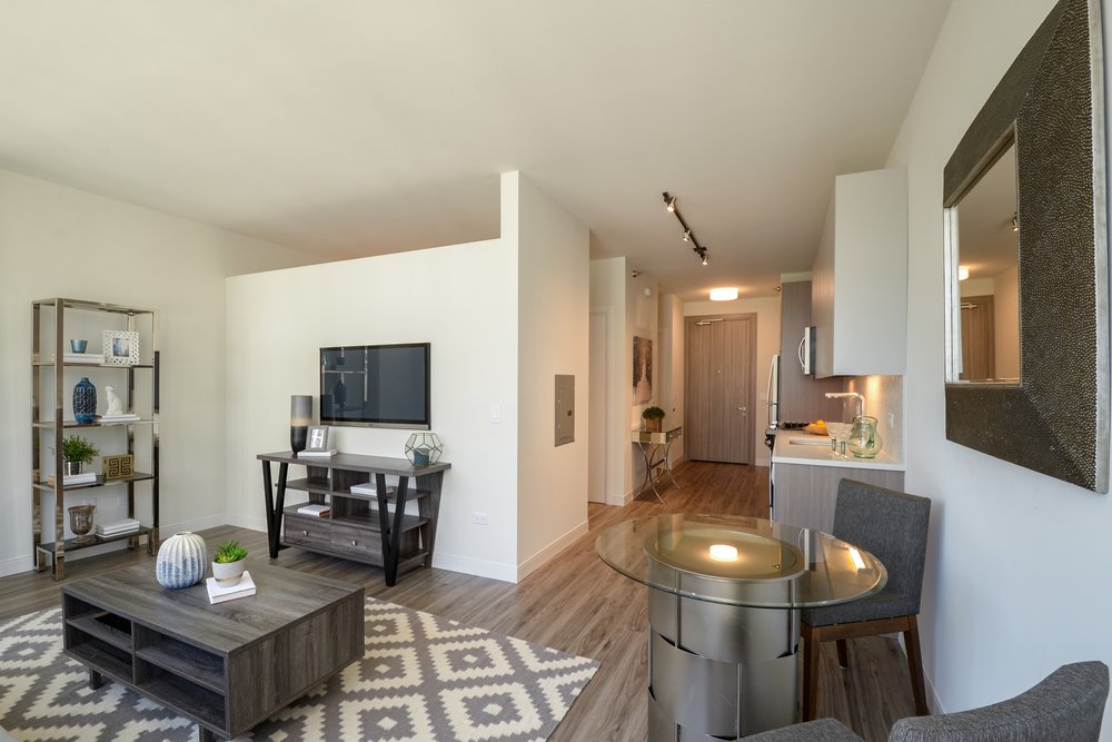 Modern apartment interior with a living area featuring a TV, shelves, and coffee table. Dining area includes a round table with two chairs. Kitchen visible in the background. Neutral color palette.