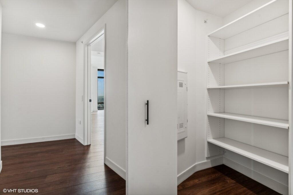 Hallway with dark wood flooring, an open door leading to a sunlit room, and a white closet with empty shelves.