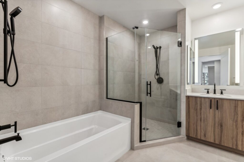 Modern bathroom with beige tiles, glass-enclosed shower, and a white bathtub. Double sink vanity with wooden cabinets and black fixtures is on the right.