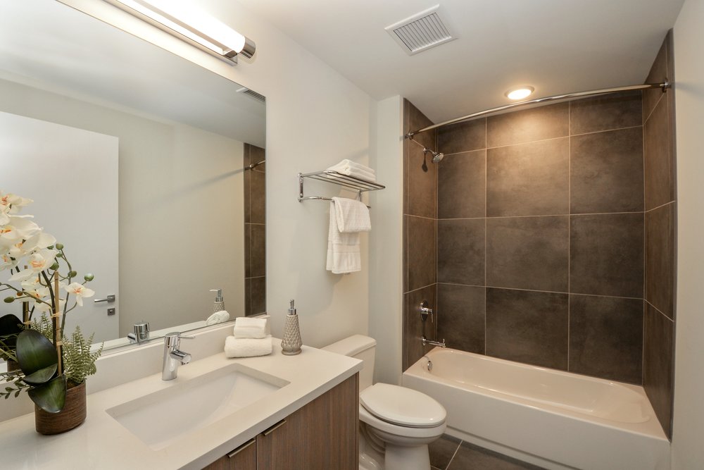 Modern bathroom with a sink, toilet, and bathtub with dark tiles. Mirror above the sink, with a towel rack and toiletries. Orchids and a plant are on the sink counter for decoration.