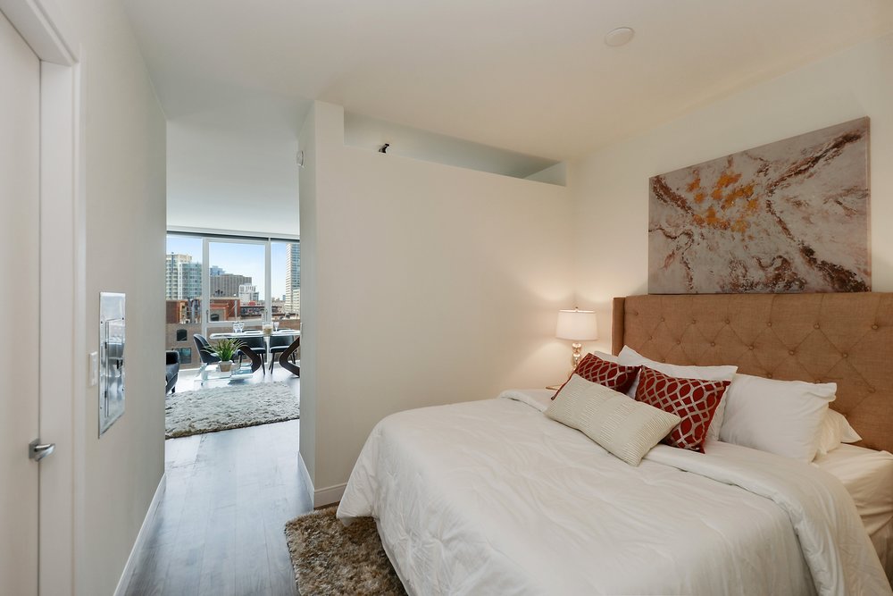 Bedroom with a large bed, cream and red pillows, wall art, and a nightstand lamp. Open door reveals a city skyline through living room windows in the background.