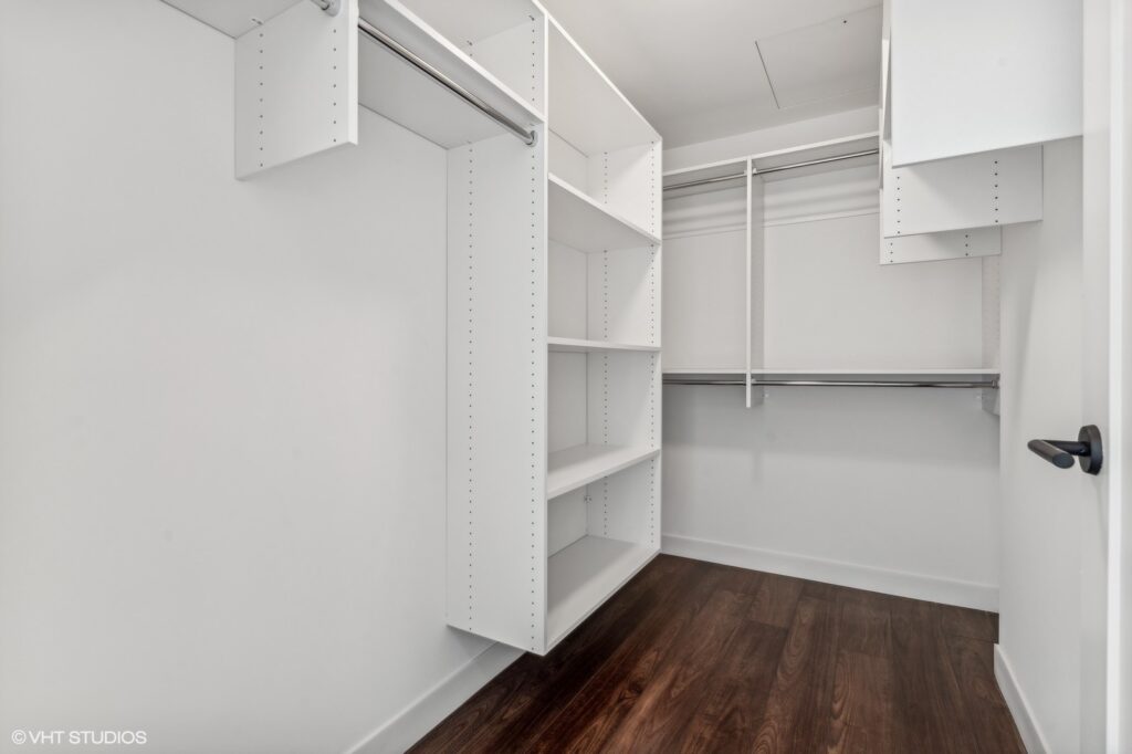 Spacious walk-in closet with white built-in shelves and clothing rods, featuring dark wood flooring and a white door.