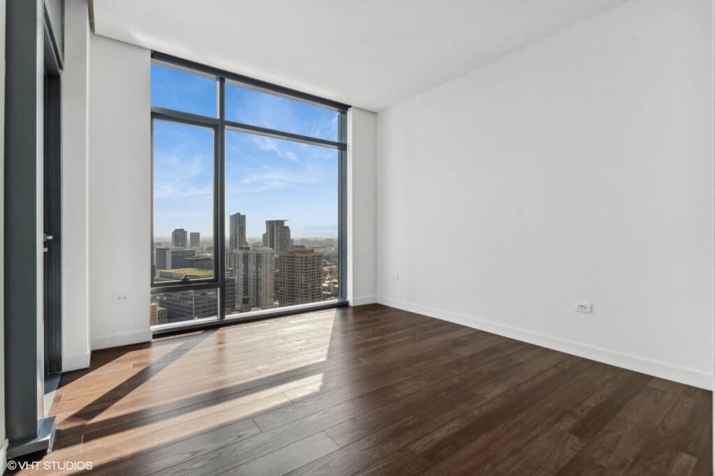 Empty room with wooden floor and large window offering a cityscape view. Walls are painted white, and there's a clear blue sky outside.
