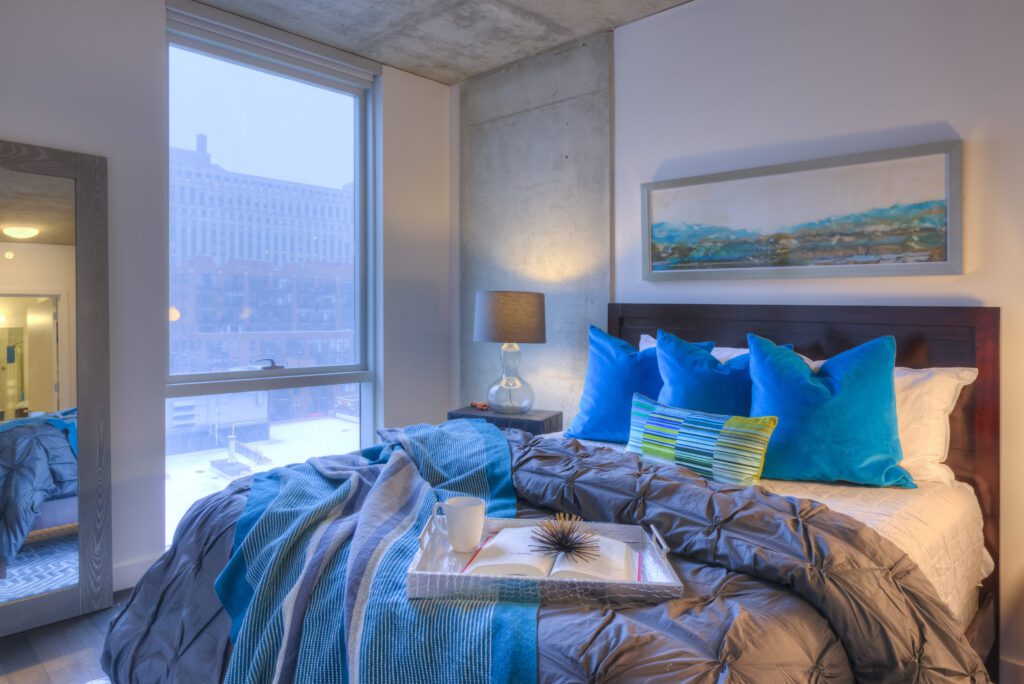 Modern bedroom with a large window showing a city view. The bed has blue and green pillows, a cozy blanket, and a tray with a mug. A lamp and abstract painting add to the decor.