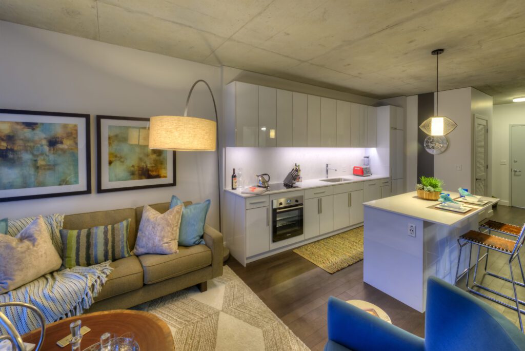Modern open-concept living room and kitchen with a beige couch, colorful throw pillows, two abstract paintings, white cabinets, and a small island. Warm lighting creates a cozy ambiance.