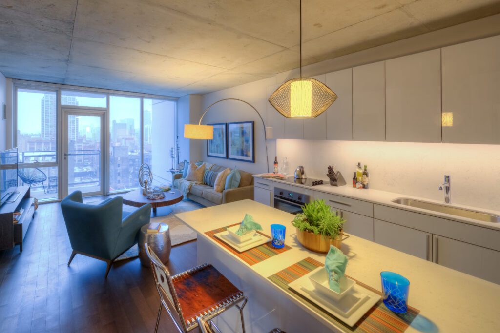 Modern open-plan apartment with a kitchen island, teal armchairs, and a couch. Large windows provide a city view. Pendant lighting and minimalistic decor are featured.