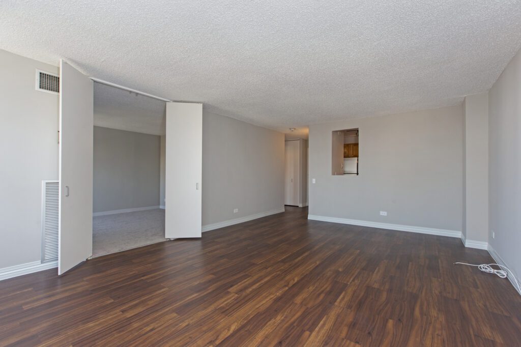 Empty room with beige walls, wooden floor, and white baseboards. Open door reveals a carpeted adjacent room. Small rectangular opening in the wall leads to a view of a kitchen area.