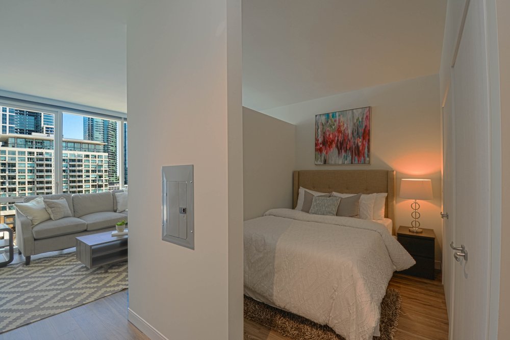 A modern apartment interior showing a bedroom with a bed and a lamp, adjacent to a living area with a sofa and coffee table. Large windows reveal a cityscape outside.