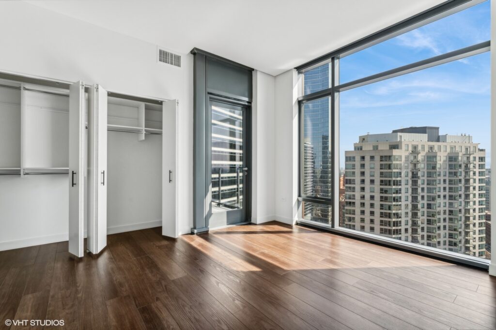 A bright, empty modern room with wooden floors, large windows, an open closet, and a cityscape view.