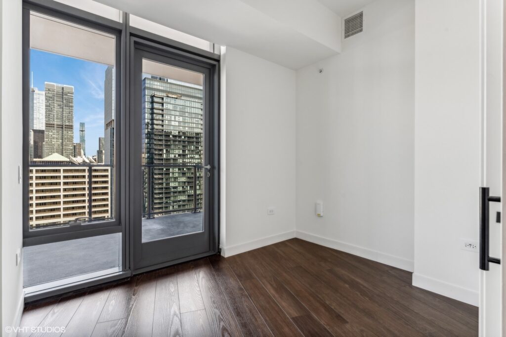 Empty room with dark wood flooring, floor-to-ceiling windows, and a glass door leading to a balcony. Cityscape with tall buildings visible outside. White walls and ceiling.