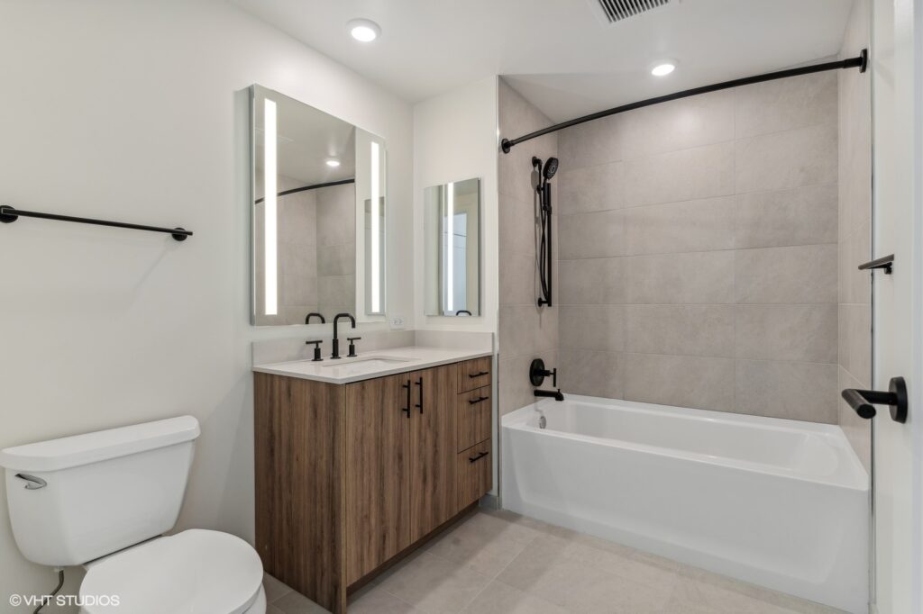 Modern bathroom with a bathtub, tile walls, a wooden vanity with double sinks, round mirrors, and black fixtures. Ceiling lights illuminate the space.