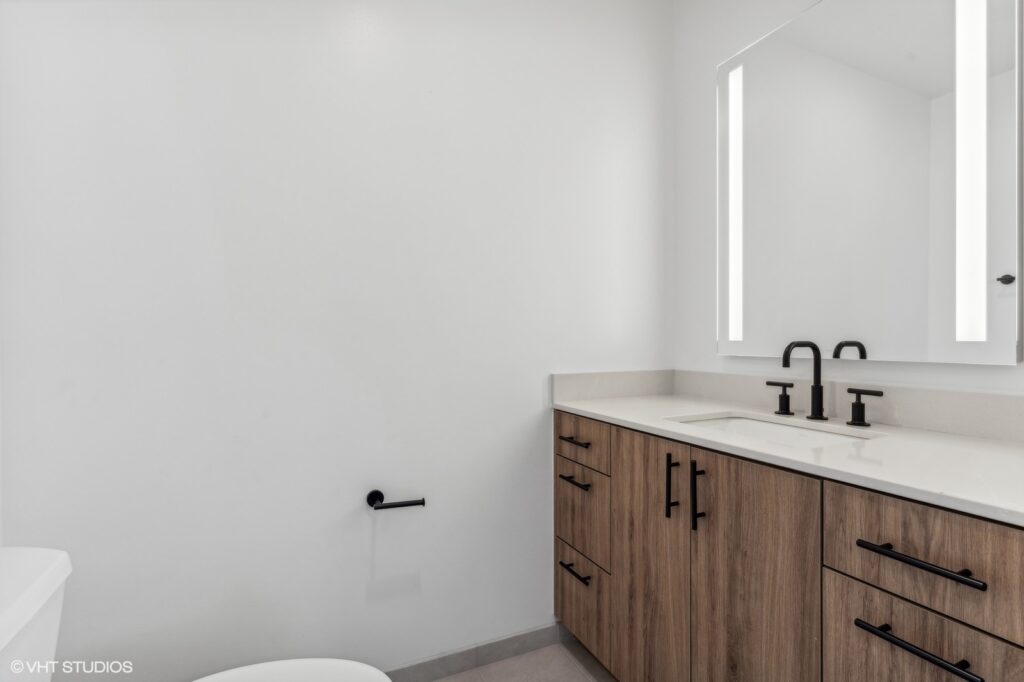 Modern bathroom with wooden vanity, rectangular mirror with integrated lighting, black faucet and handles, white walls, and a toilet.