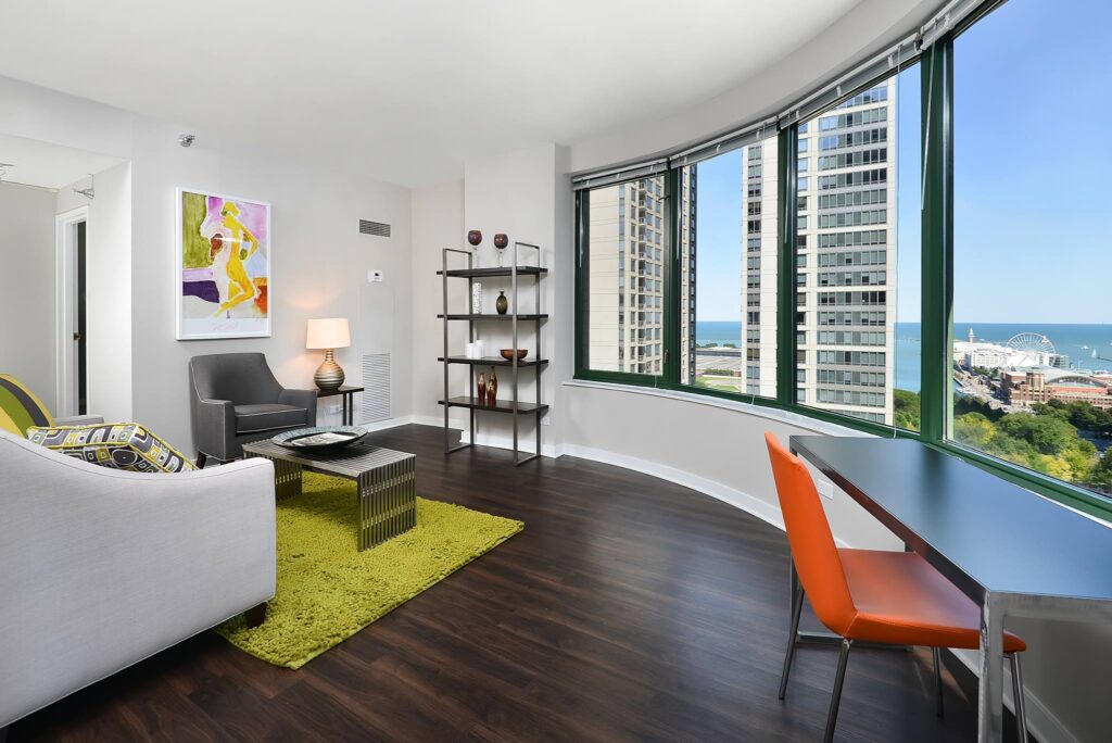 Modern living room with a white couch, gray chair, green rug, and orange chair by a desk. Large windows offer a city and waterfront view. Art hangs on the wall, and shelves hold decorative items.